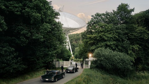 Radioteleskop, vor einem Tor Autos und Männer in schwarzen Anzügen