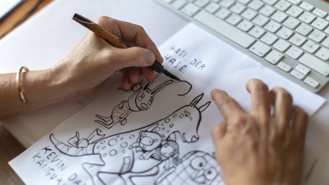 Illustratorin Kat Meschnik sitzt an einem Tisch, dabei zeichnet sie mit Tusche. 