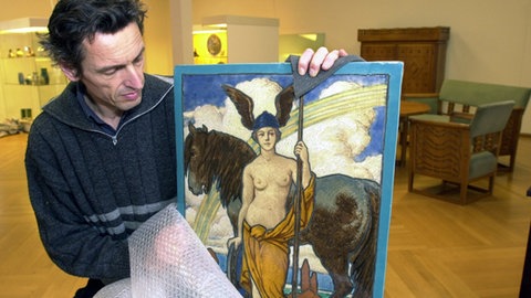 Mann packt eine Keramiktafel im Jugendstil aus, auf der eine „Minerva" abgebildet ist.