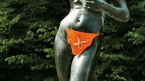 Eine Bronzestatue eines nackten Frauenkörpers. Der Intimbereich der Figur wurde mit einem orangenem Lendentuch umhäkelt.
