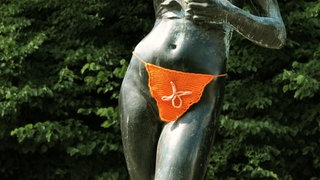 Eine Bronzestatue eines nackten Frauenkörpers. Der Intimbereich der Figur wurde mit einem orangenem Lendentuch umhäkelt.