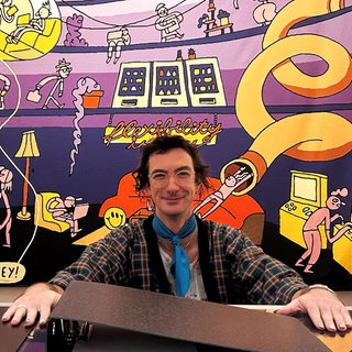 Comickünstler*in Dominik Wendland sitzt an einem überdimensionierten Schreibtisch mit riesigem Aktenordner und gewaltiger Kaffeetasse