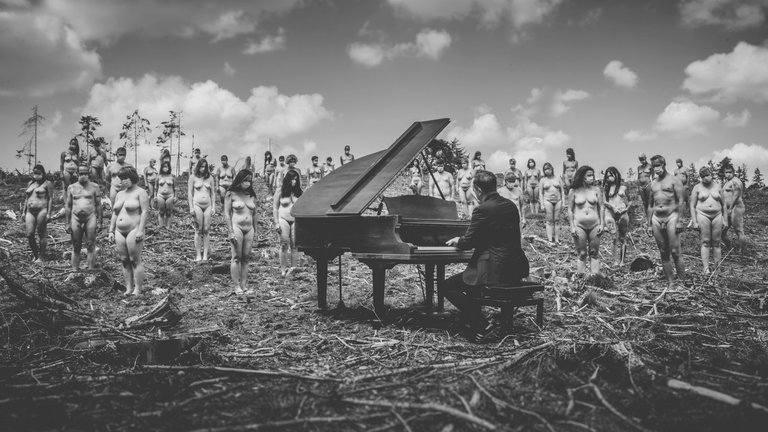 Fotowerk von Andy Reiner: Ein Pianist spielt im Freien an einem Konzertflügel, hinter ihm stehen viele unbekleidete Frauen und Männer.