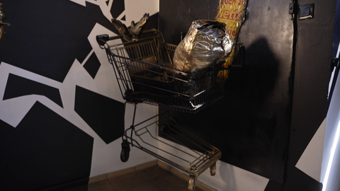 Ein Einkaufswagen mit einer gold lackiertes Einkaufstüte hängt an der Wand. Ein Kunstwerk von Ralf Blümlein. 