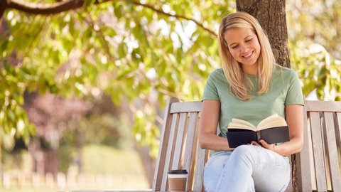 Eine Frau sitzt mit einem Buch auf einer Parkbank. Symbolbild für lesen, Romane und Buchtipps 2025.