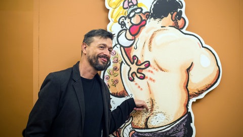 Ralf König deutet auf die Pofalte einer seiner Comic-Figuren