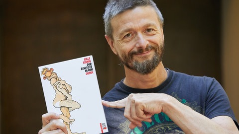 Ralf König hält eine Ausgabe seines Comics „Der bewegte Mann“ hoch.