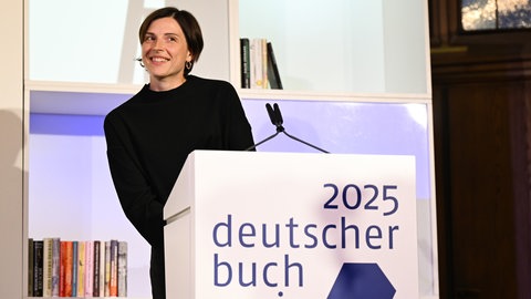 Die Schweizer Autorin Dorothee Elmiger steht lächelnd hinter einem Pult mit dem  Logo des Deutschen Buchpreises