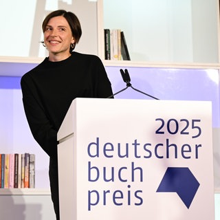 Die Schweizer Autorin Dorothee Elmiger steht lächelnd hinter einem Pult mit dem  Logo des Deutschen Buchpreises