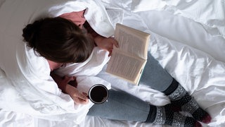 Eine Frau sitzt mit einer Tasse Tee auf einem Bett und liest ein Buch.