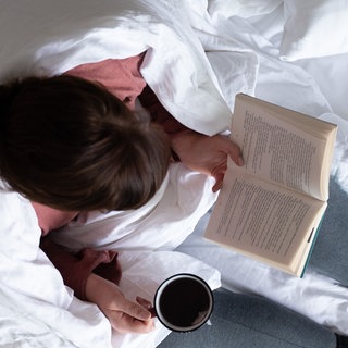Eine Frau sitzt mit einer Tasse Tee auf einem Bett und liest ein Buch.
