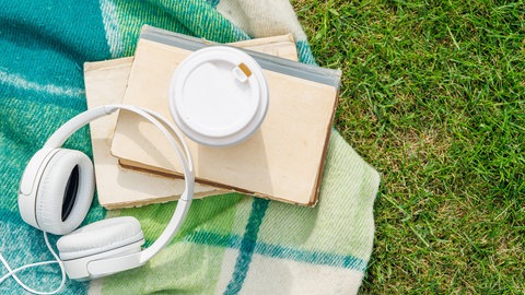 Bücher, Kopfhörer und Kaffee liegen auf einer Picknickdecke in einer Wiese