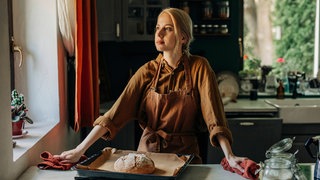 Eine blonde Frau mit Schürze steht in der Küche, vor ihr auf dem Tisch liegt ein selbstgebackenes Brot. Symbolbild für eine "Tradwife".