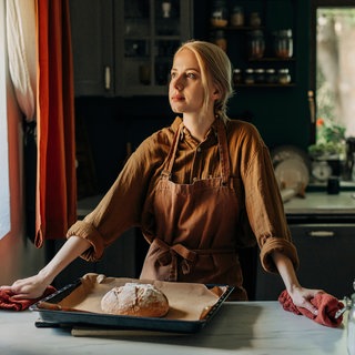 Eine blonde Frau mit Schürze steht in der Küche, vor ihr auf dem Tisch liegt ein selbstgebackenes Brot. Symbolbild für eine "Tradwife".