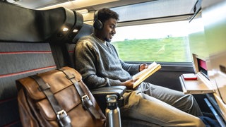 Junger schwarzer Mann, der in einem modernen Zug reist, Kopfhörer trägt und ein Buch liest.