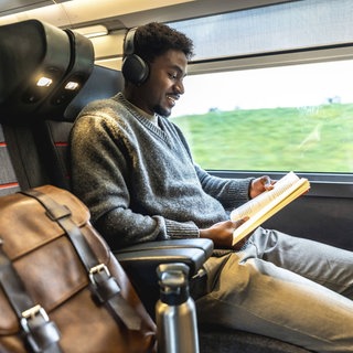 Junger schwarzer Mann, der in einem modernen Zug reist, Kopfhörer trägt und ein Buch liest.