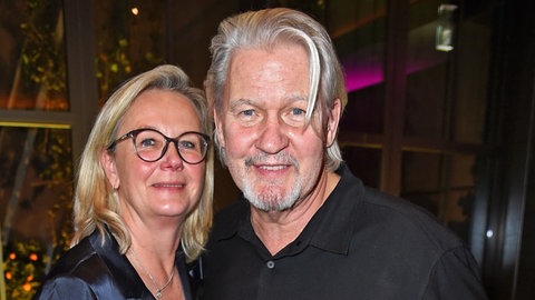 Johnny Logan und seine Partnerin Tanja Surmann halten sich im Arm und lächeln. Sie hat blondes Haar und trägt ein schwarze Brille.