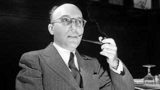 Der Komponist Kurt Weill sitzt an einem Tisch und raucht eine Pfeife.