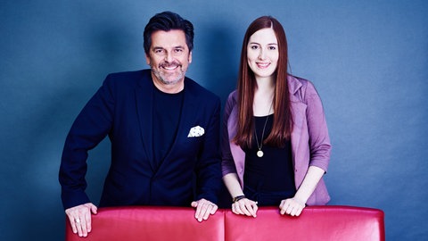 Schlagersänger Thomas Anders steht mit Musikredakteurin Andrea Mayer hinter einem roten Sofa. Beide tragen schicke Blazer und lächeln nach dem Interview in die Kamera.