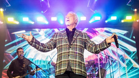 Tom Jones steht auf eine hellbeleuchteten Bühne und hebt die Arme und hat die Augen geschlossen