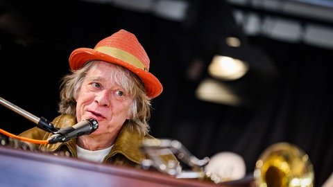 Helge Schneider mit rotem Hut sitzt am Klavier und spricht in ein Mikrofon