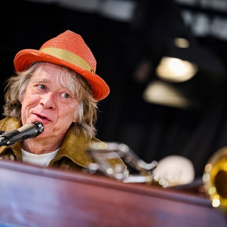 Helge Schneider mit rotem Hut sitzt am Klavier und spricht in ein Mikrofon