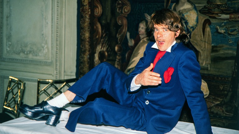 Helge Schneider im blauen Anzug und roter Krawatte in komischer Pose auf Tisch