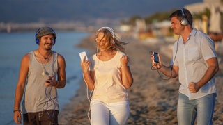 Drei junge Leute genießen einen Spaziergang am Strand und hören dabei Musik.