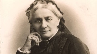 Fotografie der betagten Clara Schumann von Wilhelm Dreesen (1899). Schumann blickt direkt in die Kamera, das Kinn leicht auf die rechte Hand gestützt. Sie trägt ein schwarzes Kleid und eine schwarze Haube.