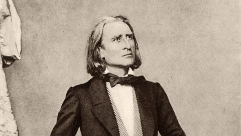 Schwarz-Weiß-Porträt des jungen Franz Liszt. Der Komponist steht in einem Anzug aufrecht, den Arm auf eine Stuhllehne angelegt.