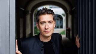 Halbtotale von Pianist Yevgeny Sudbin im Gang stehend und direkt in die Kamera schauend