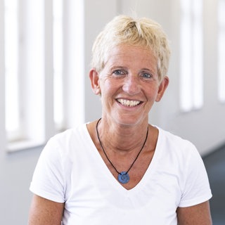 Susanne Benda - blondes kurzes Haar, weißes T-Shirt und eine Halskette mit Anhänger