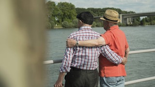 Symbolbild zum Tag der Freundschaft: Zwei ältere Männer stehen Arm in Arm an einem Flussufer
