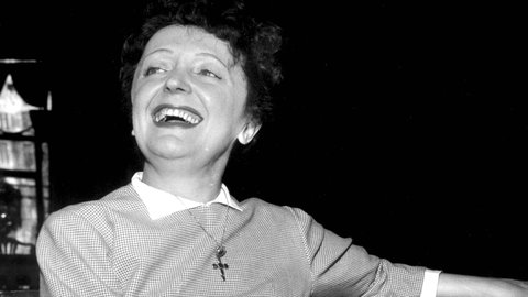 Die französische Sängerin und "Spatz von Paris" Édith Piaf sitzt auf einem Stuhl an einem Tisch, hat einen Arm auf den Tisch gelegt, schaut in die Ferne und lacht.