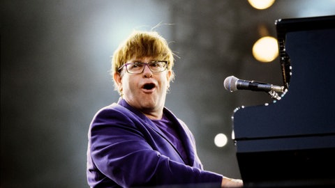Quiz über Stars: Der britische Sänger Elton John sitzt am Klavier und singt in einem lila Sakko.
