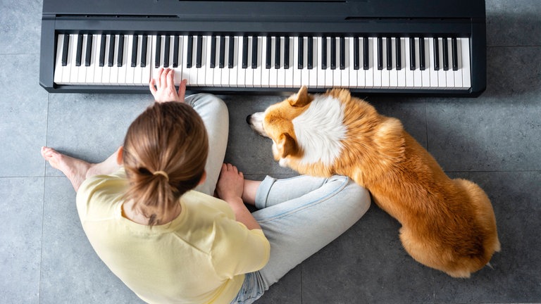Frau mittleren Alters sitzt auf dem Boden und übt Klavierspielen zu Hause. Ihr Hund liegt daneben.