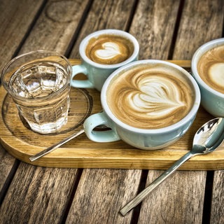 Drei Kaffeetassen mit Herz-Latte-Art-Muster auf einem Holztablett, begleitet von einem Glas Wasser