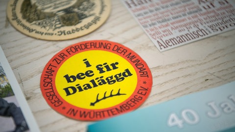 Aufkleber mit der Aufschrift "i bee fir Dialäggd": Ob Sprache oder Dialekt unterscheidet sich durch Kriterien wie Standardisierung, vereinheitliche Schreibung, gegenseitige Verständlichkeit und das Dialektkontinuum