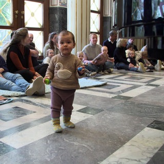 Babkonzert in Stuttgarter Marmorsaal: Ein Baby steht in der MItte und tanzt, während Eltern am Rand sitzen und zuschauen. 