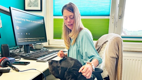 SWR1 Neuanfang: Heike sitzt am Schreibtisch, vor ihr ein Computer-Bildschirm mit besonders großen Buchstaben. "Neuanfang: Wenn es anders kommt im Leben" steht dort. Neben Heike sitzt Blindenführhund Zembo und lässt sich streicheln.