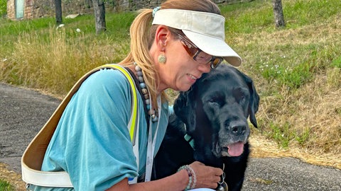 SWR1 Neuanfang: Heike - eine blonde Frau mit Pferdeschwanz - umarmt einen Hund mit schwarzem Fell. Zembo ist ihr Blindenführhund.