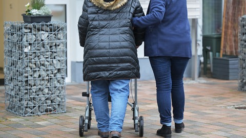 Eine Seniorin geht am Arm einer Pflegedienst-Mitarbeiterin zu ihrer Wohnung