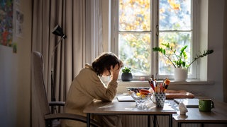 Prokrastination: Erschöpfte Frau mit beruflichem Burnout sitzt am Tisch, hält den Kopf in den Händen und versucht, mit der Arbeit zu beginnen. Eine neue Studie hat nun eine mögliche Erklärung gefunden, wieso unser Startknopf im Gehirn manchmal klemmt.