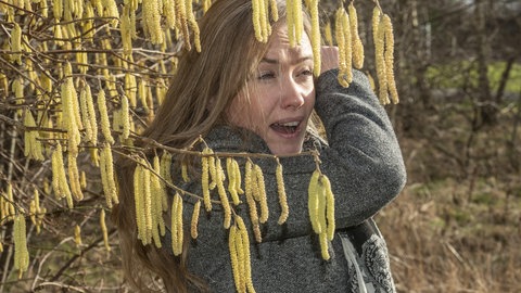 Frau steht im Frühling an blühendem Haselnuss-Strauch und hat mit Allergie zu kämpfen.