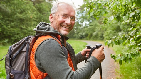 Rente und Ruhestand gut planen: Ein älterer Mann läuft mit Wanderstöcken durch die Natur und dreht sich lächelnd um.