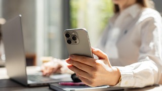 Frau im weißen Hemd beim Multitasking mit Smartphone und Laptop in einem hellen, modernen Büro. | Warum wir Produktivität bei der Arbeit oft vortäuschen