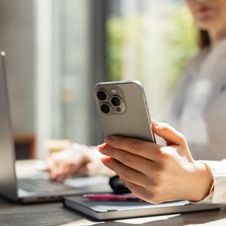 Frau im weißen Hemd beim Multitasking mit Smartphone und Laptop in einem hellen, modernen Büro. | Warum wir Produktivität bei der Arbeit oft vortäuschen