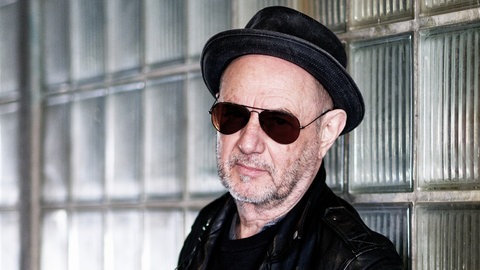 Stefan Waggershausen mit Hut und Sonnenbrille lehnt sich gegen eine Glaswand