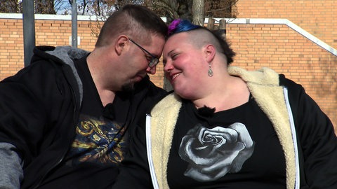 Lukas und Kathi aus Ostfildern sitzen auf einer Bank im Freien und lehnen verliebt die Köpfe aneinander.