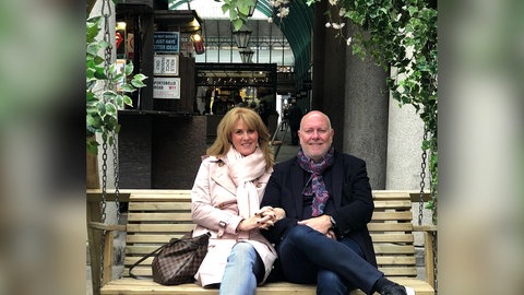 Kim und Christian sind seit 36 Jahren ein Paar. Sie sitzen nebeneinander auf einer Holzbank.